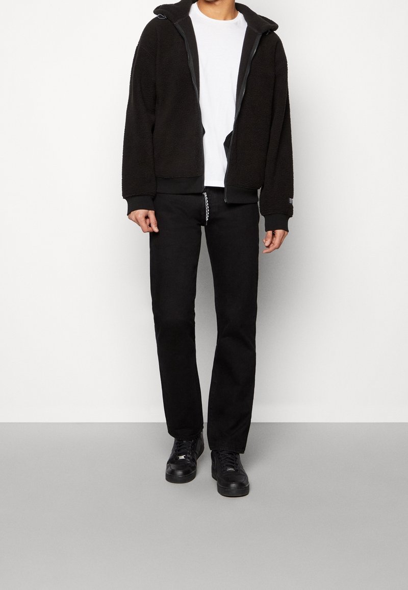 Chaqueta de forro polar negra con capucha, cierre frontal y puños acanalados. Llevada sobre una camiseta blanca y pantalones negros. Zapatillas negras con superficie texturizada.