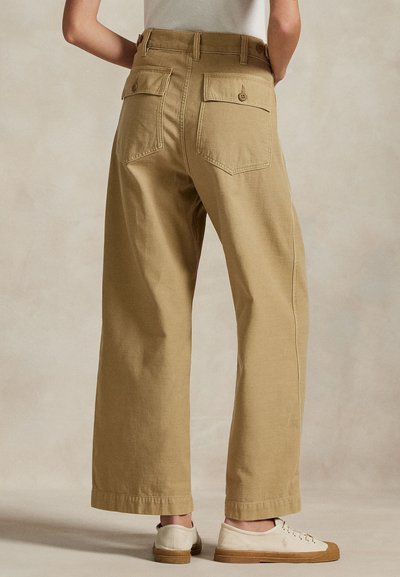 Μπεζ παντελόνια με φαρδιά μπατζάκια και πίσω τσέπες με κουμπιά, φορεμένα με λευκά αθλητικά παπούτσια, απεικονιζόμενα από πίσω σε ένα άτομο που στέκεται.