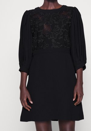 Musta mekko, jossa on teksturoitu pitsirintamikko kukkakuvioilla, pussimaiset hihat ja vartalonmyötäinen siluetti. Sileä hame, jossa hillittyjä viivadetaljeja.