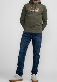Oliwkowa bluza z kapturem z pomarańczowo-białym logotypem, zestawiona z ciemnoniebieskimi dżinsami i beżowymi trampkami.