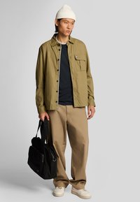 Giacca camicia leggera beige con taschino, indossata sopra una maglietta scura, abbinata a pantaloni khaki chiari e sneakers bianche, mentre tiene una borsa nera.
