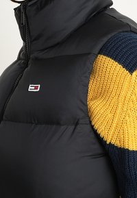 Czarna kamizelka puchowa z wysokim kołnierzem, z logo. Widoczna jest połowa dzianinowego swetra z żółtymi i granatowymi paskami obok.