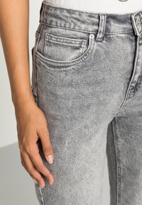 Grijze denim jeans met een vervaagde textuur, vijf-pocket ontwerp en zichtbare stiksels. Hand rustend op het bovenbeen, waardoor de details zichtbaar zijn.