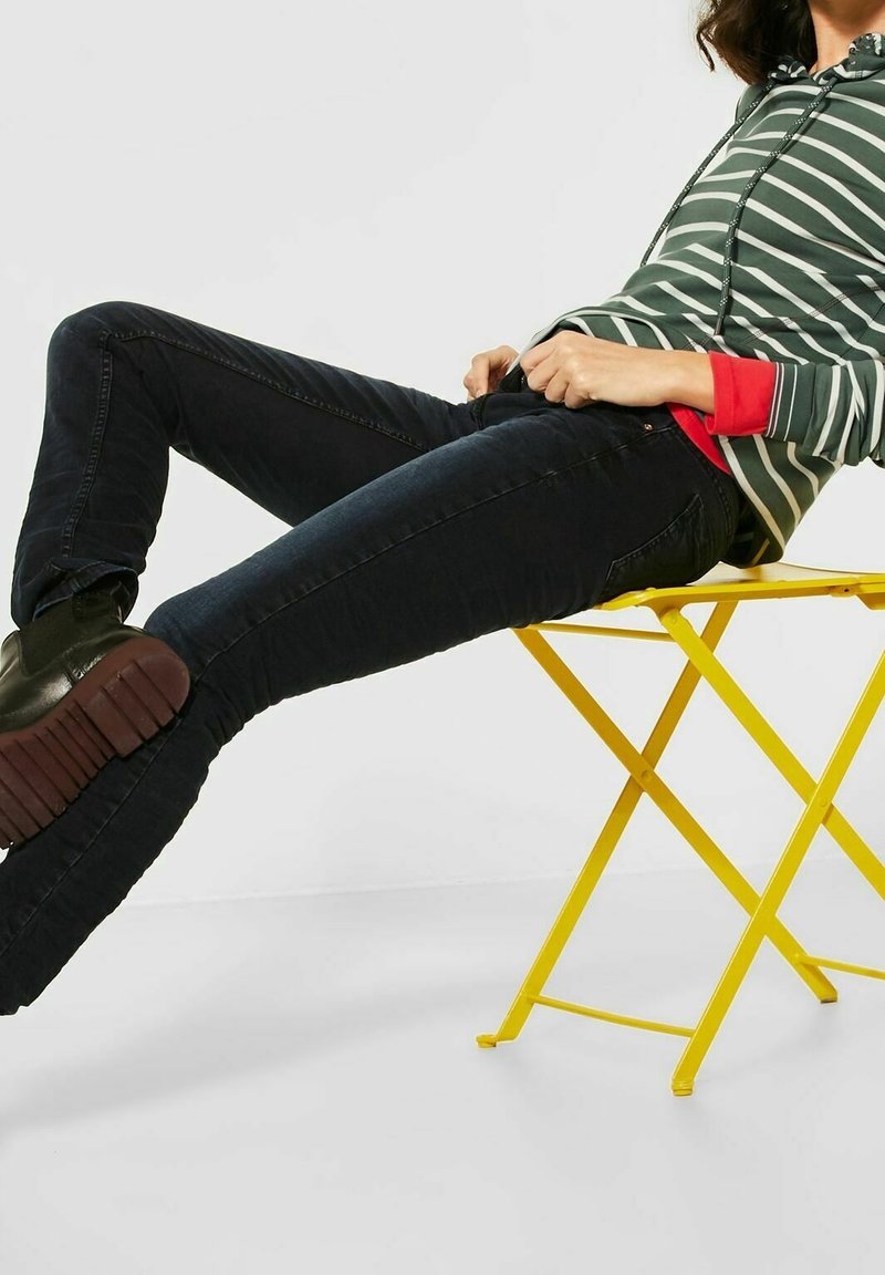 Persona che indossa jeans neri, una felpa a righe verde e stivali neri, sdraiata su una sedia pieghevole metallica gialla su uno sfondo bianco.