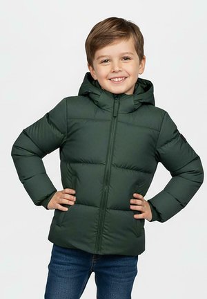 Lachende jonge jongen staand met handen in de zij, draag een donkergroene donsjas met capuchon en blauwe jeans tegen een effen achtergrond.