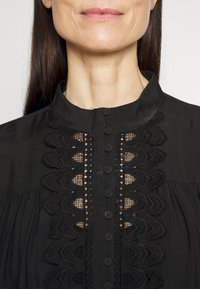 Μαύρη μπλούζα με ψηλό γιακά, διακοσμημένη με δαντελωτές κοπές και κυματιστές λεπτομέρειες. Κλείσιμο με κουμπιά μπροστά με μικρά μαύρα κουμπιά. Λείος υφασματικός ιστός.