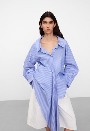 Femme aux longs cheveux foncés portant une chemise oversize rayée bleue et blanche et une jupe blanche, debout devant un fond uni.