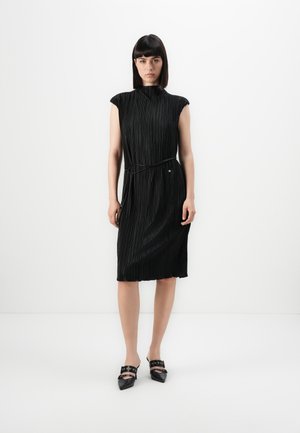 Woman with straight dark hair wearing a sleeveless black pleated midi dress with a thin belt, standing against a plain white background.