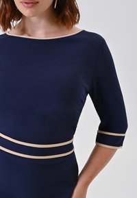 Frau trägt ein navyblaues Kleid mit beiger Einfassung am Ausschnitt, in der Taille und an den Ärmeln, vor einem schlichten hellen Hintergrund.