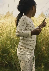 Barn med mörkt hår i en krämfärgad blommig outfit, håller i höga gräs strå, står i ett solbelyst fält av vilda gräs.