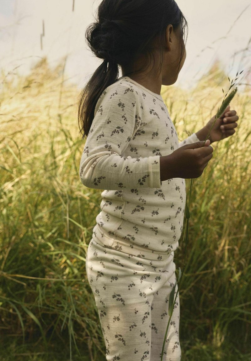 Barn med mörkt hår i en krämfärgad blommig outfit, håller i höga gräs strå, står i ett solbelyst fält av vilda gräs.