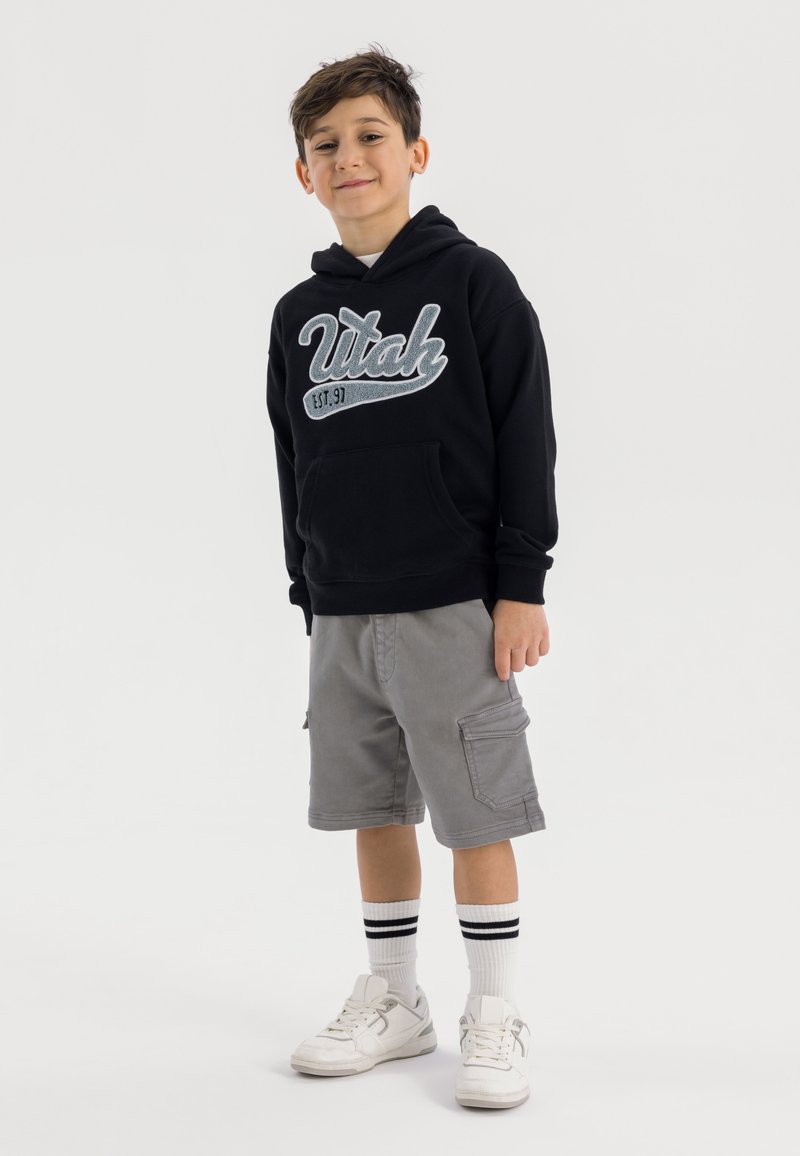 Svart luvtröja med "Utah" grafik, grå cargoshorts, vita strumpor med svarta ränder och vita sneakers. Barn står självsäkert.