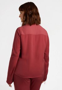 Blusa a maniche lunghe di un rosso intenso, caratterizzata da una trama liscia e una parte superiore semi-trasparente. Progettata con un taglio dritto e dettagli minimi.