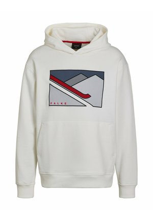 Kobieta stojąca w białej bluzie z kapturem z grafiką gór i narciarza, czarnych legginsach i białych trampkach na jednolitym tle.