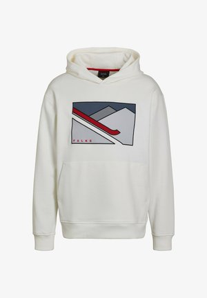 Vrouw staand, gekleed in een witte hoodie met berg- en skigrafiek, zwarte legging en witte sneakers tegen een effen achtergrond.