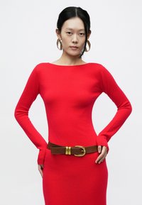 Czerwona sukienka z długim rękawem o dopasowanym kroju i prążkowanej fakturze. Połączona z brązowym paskiem z złotą klamrą i dużymi kolczykami kołami.