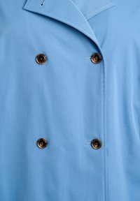 Světlounce modrý dvouřadý kabát s klopovým límcem, zdobený dvěma tmavě hnědými knoflíky a hladkou texturou látky.