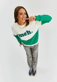 Zelená a bílá mikina s nápisem "meh.", šedé kalhoty a vícebarevné tenisky. Měkká tkanina, volný střih a ležérní design.