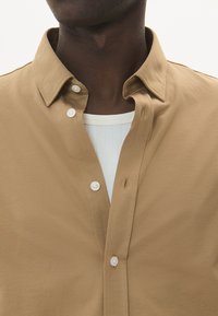 Hnedá košeľa na gombíky so štruktúrovaným golierom, s bielymi gombíkmi a hladkým, textúrovaným materiálom. Nosená cez biely rebrovaný tielko.