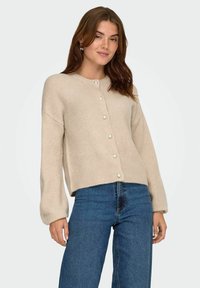 Beige Strickjacke mit Rundhalsausschnitt und langen Ärmeln, versehen mit Perlenknöpfen auf der Vorderseite. Kombiniert mit blauen weit geschnittenen Jeans.