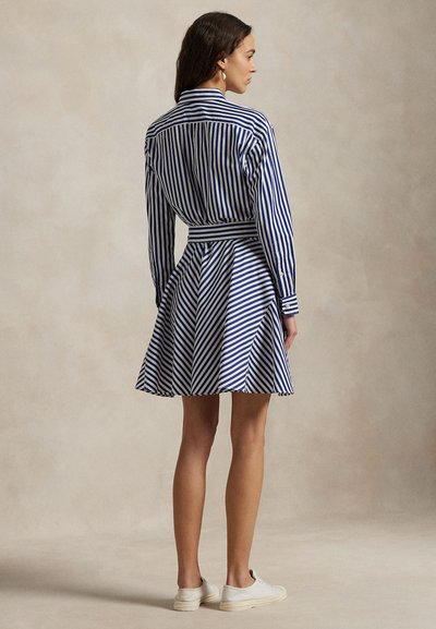 Sieviete, tērpusies zilā un baltā vertikāli svītrotā garroku kleitā ar izvērstu svārku un baltās sporta kurpēs, stāvot ar muguru pret mums.