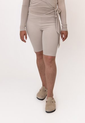 Personne portant un haut beige à manches longues avec un lien sur le côté, un short assorti longueur genou et des chaussures beige sans lacets, face à l’avant sur fond uni.