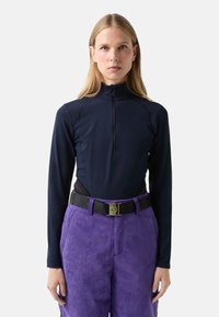 Žena s dlouhými blond vlasy má na sobě přiléhavý tmavě modrý top na zip s dlouhými rukávy a vysokým pasem fialové manšestrové kalhoty s černým opaskem.