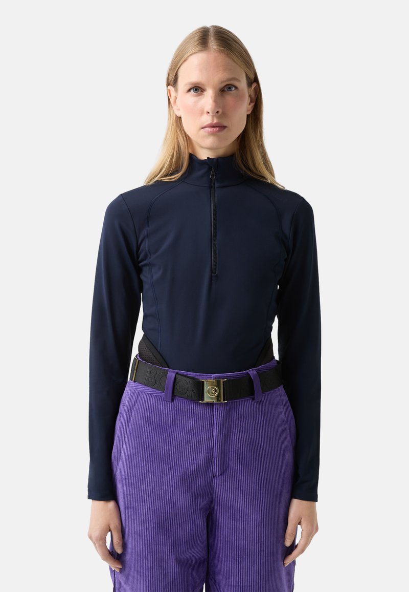 Žena s dlouhými blond vlasy má na sobě přiléhavý tmavě modrý top na zip s dlouhými rukávy a vysokým pasem fialové manšestrové kalhoty s černým opaskem.