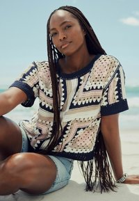Hæklet sweater i marineblå, pink, creme og beige med geometriske mønstre; parret med denimshorts. Model sidder på sand, sollys ovenfor.