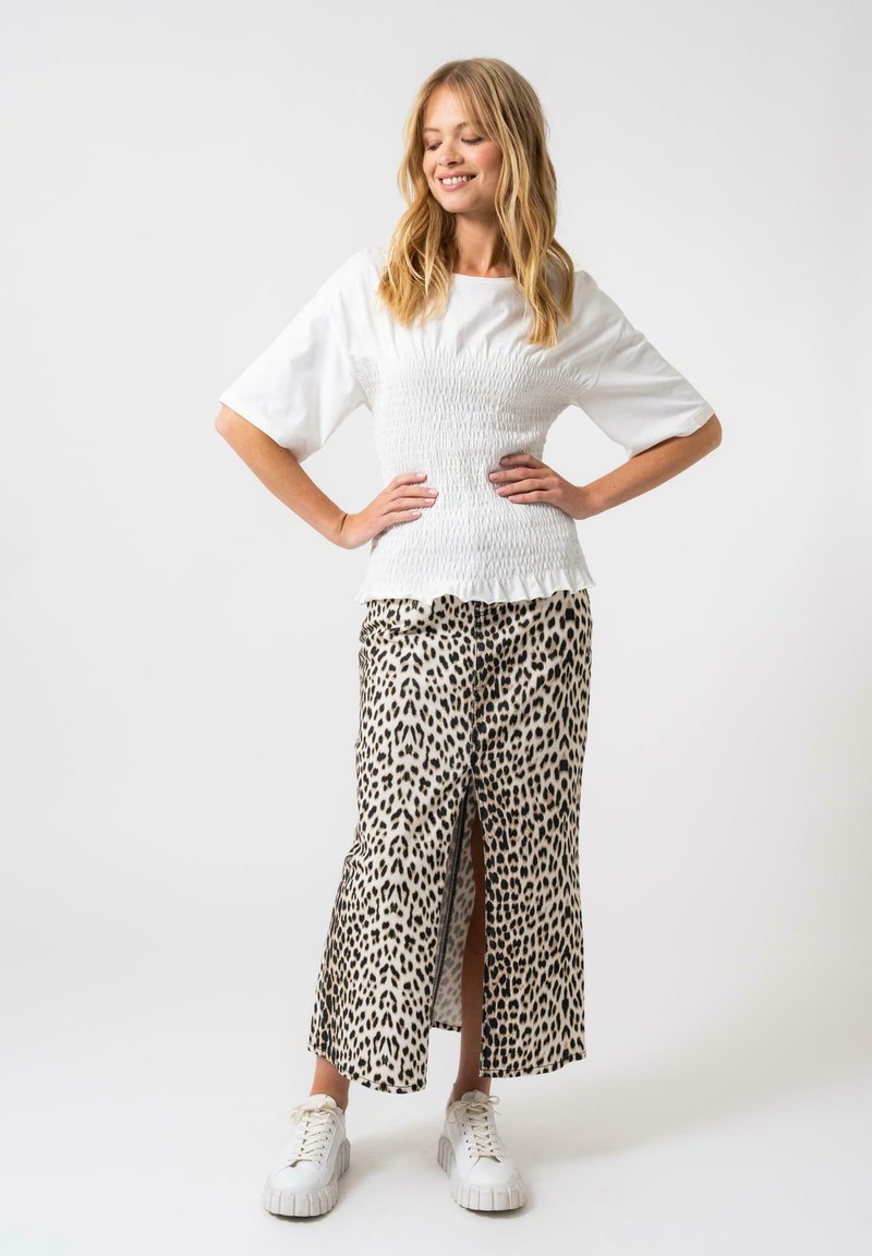 Top bianco con arricciature e maniche corte e ampie, abbinato a una gonna maxi con stampa leopardata e spacco anteriore, indossato con sneakers bianche.