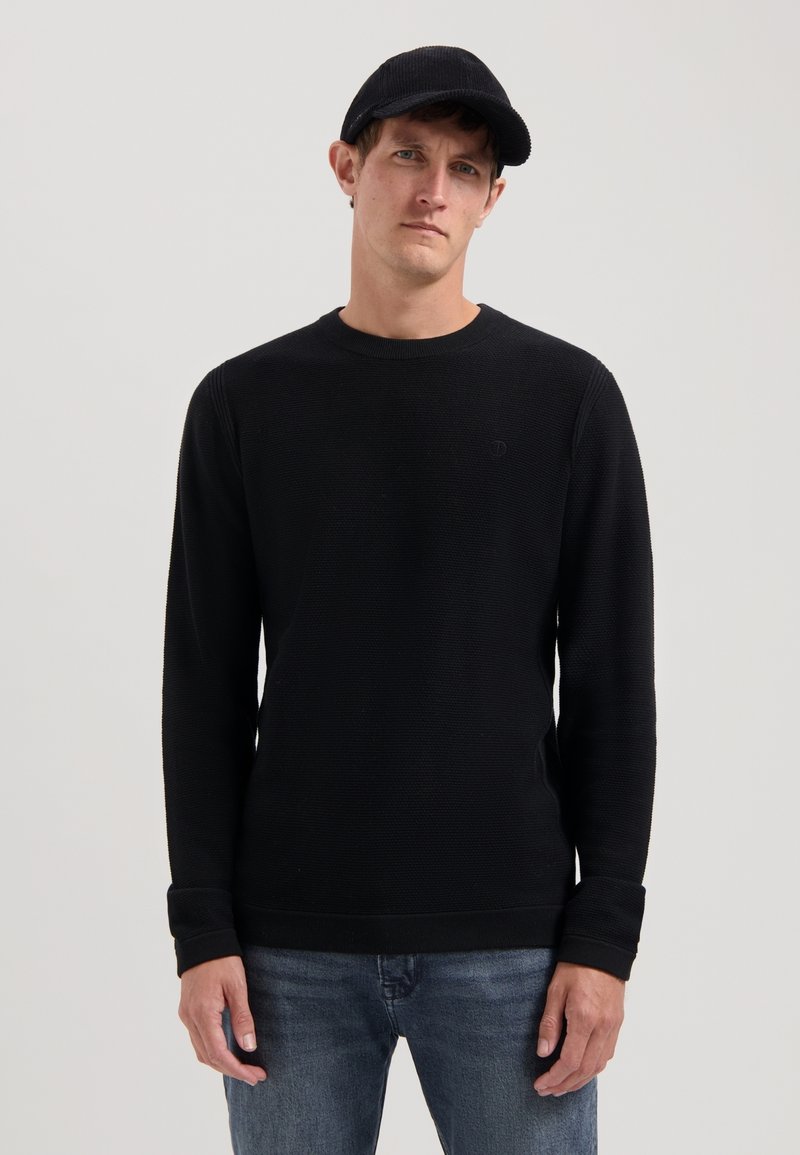 Schwarzer Strickpullover mit rundem Halsausschnitt und langen Ärmeln, ausgestattet mit gerippten Bündchen und einem dezenten Logo auf der Brust. Kombiniert mit Jeans.