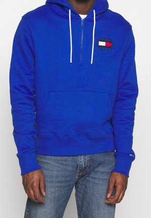 Hoodie zippé bleu en tissu doux, avec une poche avant, des lacets blancs et un patch logo rouge et blanc sur la poitrine.