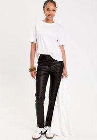 Pantalones de cuero sintético negros, camiseta de algodón blanca y chaqueta blanca en la mano. Presenta trenzas y zapatillas blancas con acentos grises y negros.