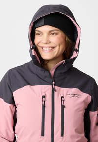 Giacca invernale rosa e grigia scura con cappuccio, dotata di due cerniere frontali, cordini regolabili e un logo sul petto. Cappello lavorato a maglia nera.