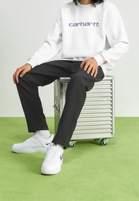Felpa grigia con logo "carhartt", pantaloni neri a coste e sneakers bianche Nike Air Force 1 su una cassa di stoccaggio metallica. Pavimento verde.