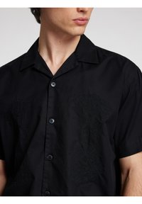 Camicia nera a maniche corte con colletto a punta, due bottoni e ricami testurizzati sul davanti. Vestibilità classica con una finitura in tessuto liscia.