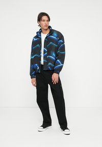 Čierna bomber bunda s modrými abstraktnými vzormi, so zipsom vpredu a elastickými manžetami. Nosená cez biely tričko a skombinovaná s čiernymi cargo nohavicami a teniskami.