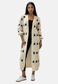 Strickstrickjacke in Creme mit langem, offenem Design und schwarzen Sternmustern; detaillierte gerippte Bündchen und Saum; getragen über einem schwarzen Oberteil.