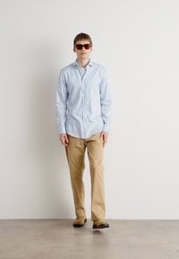 Light blue button-up shirt, khaki pants, dark sunglasses, with a relaxed fit and smooth texture. Floor is light gray, background is plain white.