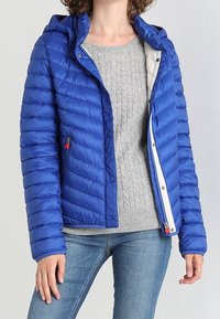 Blå pufferjakke med hætte, kviltet tekstur, lynlås foran og sidelommer. Båret over en grå strikket sweater og denim jeans.