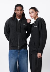 Svarte zip-up hettejakker laget av myk, teksturert stoff, med lommer foran og en trykt "Blessed" logo på brystet. Vist av to personer.
