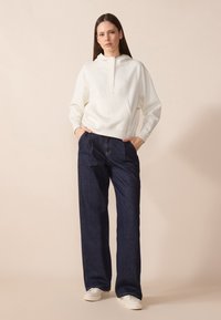Sweatshirt à capuche blanche avec une poche kangourou, associé à un jean large bleu foncé et des baskets blanches. Ensemble simple et décontracté.