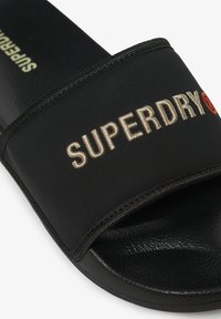 Zwarte schuivende sandaal met een dikke stoffen band waarop "SUPERDRY" in goud geborduurd is. Getextureerde rubberen zool met een gladde afwerking.