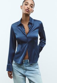 Marineblå satin langærmet skjorte med krave, knaplukning foran og let afslappet pasform, parret med lyseblå slidte jeans.