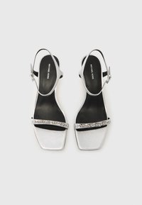 Ασημένια σανδάλια με τετράγωνη μύτη, διαθέτουν κομψό μεταλλικό φινίρισμα και μαύρες εσωτερικές σόλες. Περιλαμβάνουν διακοσμητικό λουράκι με λογότυπα.