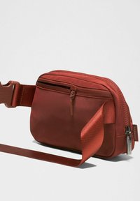 Crvena torbica preko tijela od tkanine s glatkom teksturom, zatvaranje na zip, podesivim remenom i oznakom s logotipom. Kompaktni pravokutni oblik.