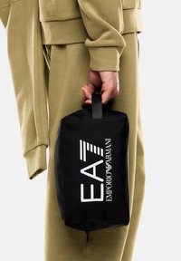 Neceser de marca negro con asa en la parte superior, que presenta el logo "EA7 EMPORIO ARMANI" en blanco, hecho de tela resistente, de forma rectangular.