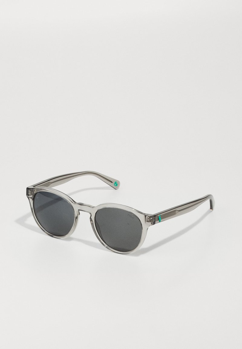 Gafas de sol con un marco gris transparente, lentes gris oscuro y brazos plateados texturizados. Presenta pequeños acentos turquesa cerca de las bisagras.