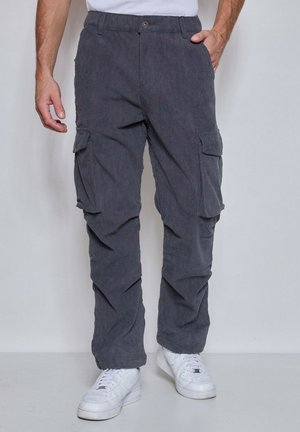 Homme portant un pantalon cargo en velours côtelé gris foncé et des baskets blanches, une main dans la poche, debout devant un fond uni.