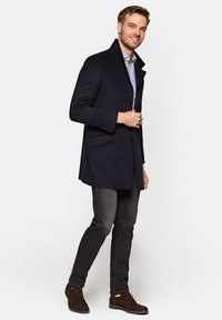 Granatowy wełniany płaszcz z wysokim kołnierzem, zapinany na pojedynczy guzik, z kieszeniami z przodu, noszony na paskowanej koszuli, w zestawieniu z ciemnymi dżinsami i brązowymi butami.